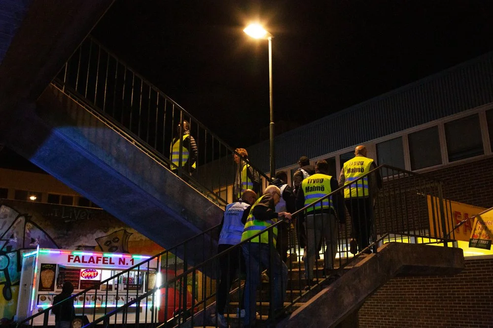 Group of people wearing reflective vests walking up an outdoor staircase at night, illuminated by a streetlamp, near a food truck and graffiti wall.
