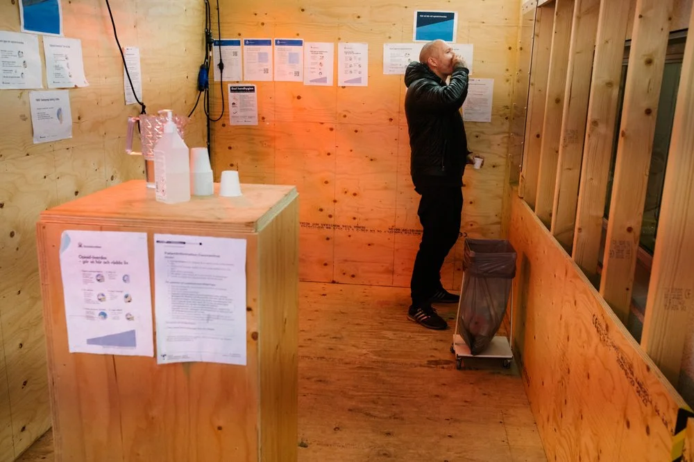 A man in a black jacket and black pants standing inside a wooden booth, drinking from a cup. The booth has wooden walls, some informational papers posted on the walls and a wooden counter with disposable cups and a bottle on it. There is a small tras