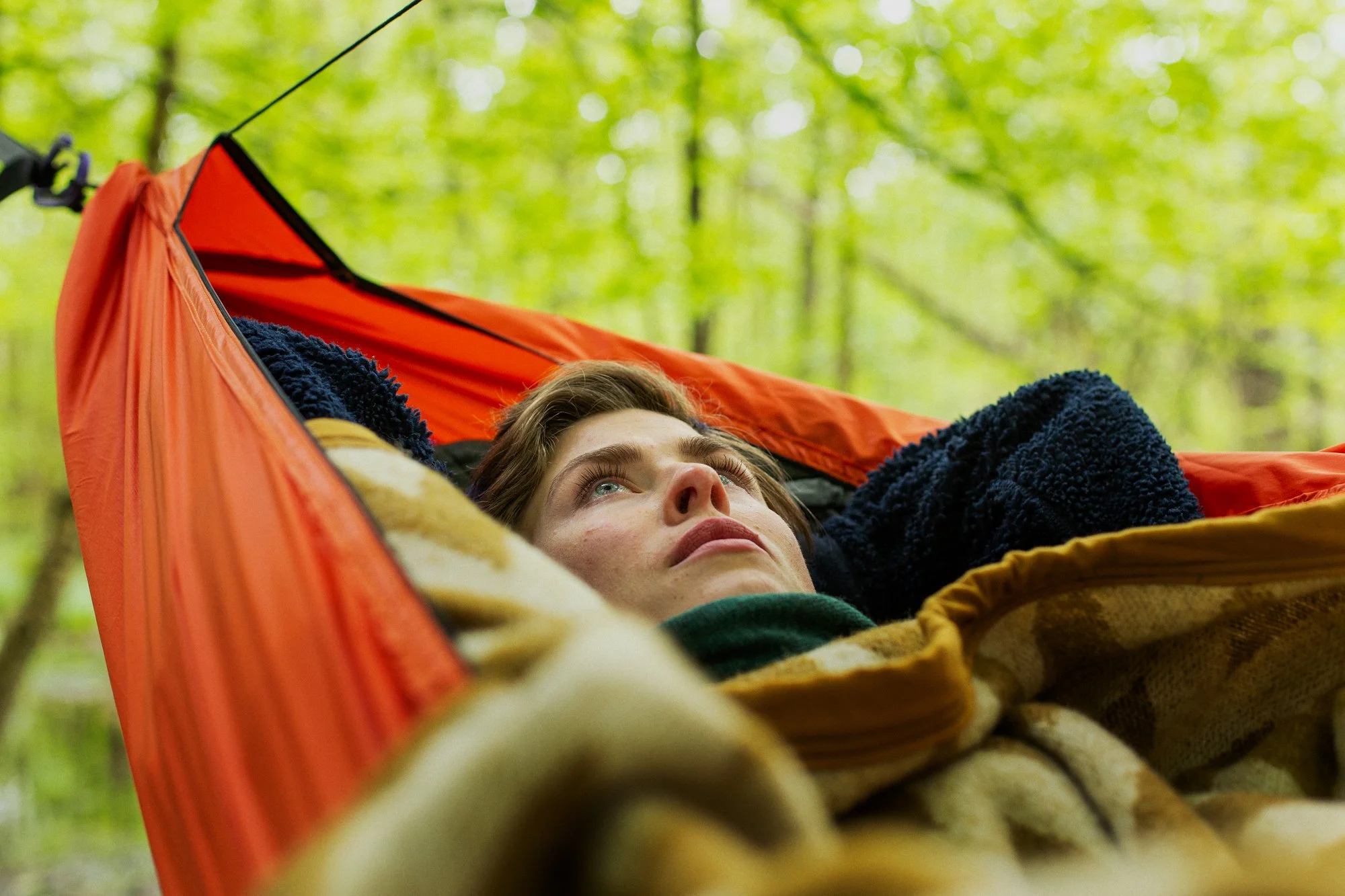 A person lying in a hammock in the woods, looking up at the sky. The hammock has a bright orange exterior and the person is covered with a patterned blanket.
