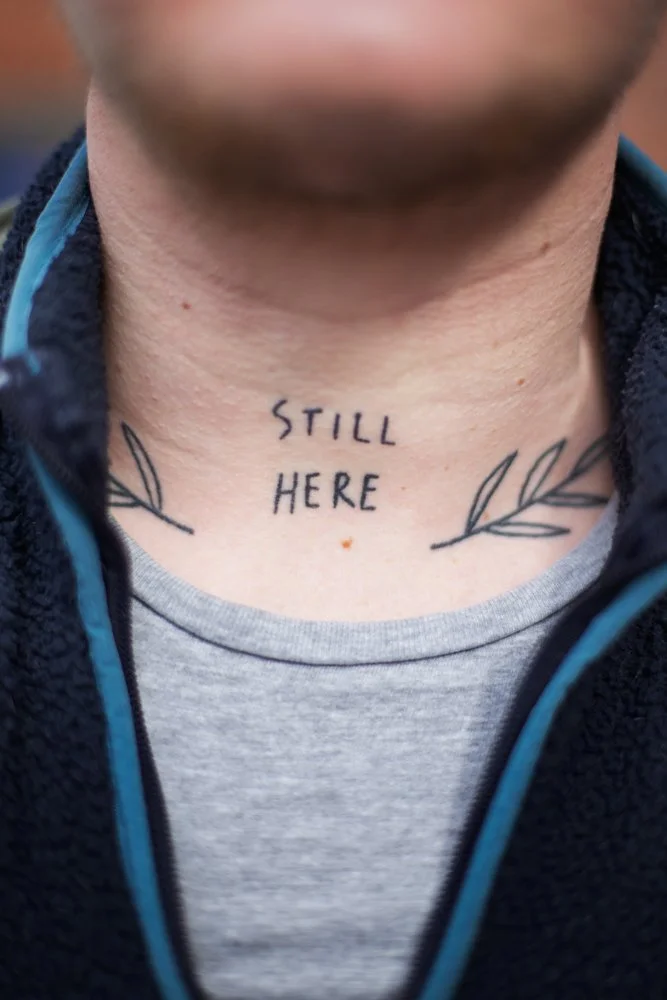 Tattoo on person's neck reading 'STILL HERE' with leaf designs on each side.