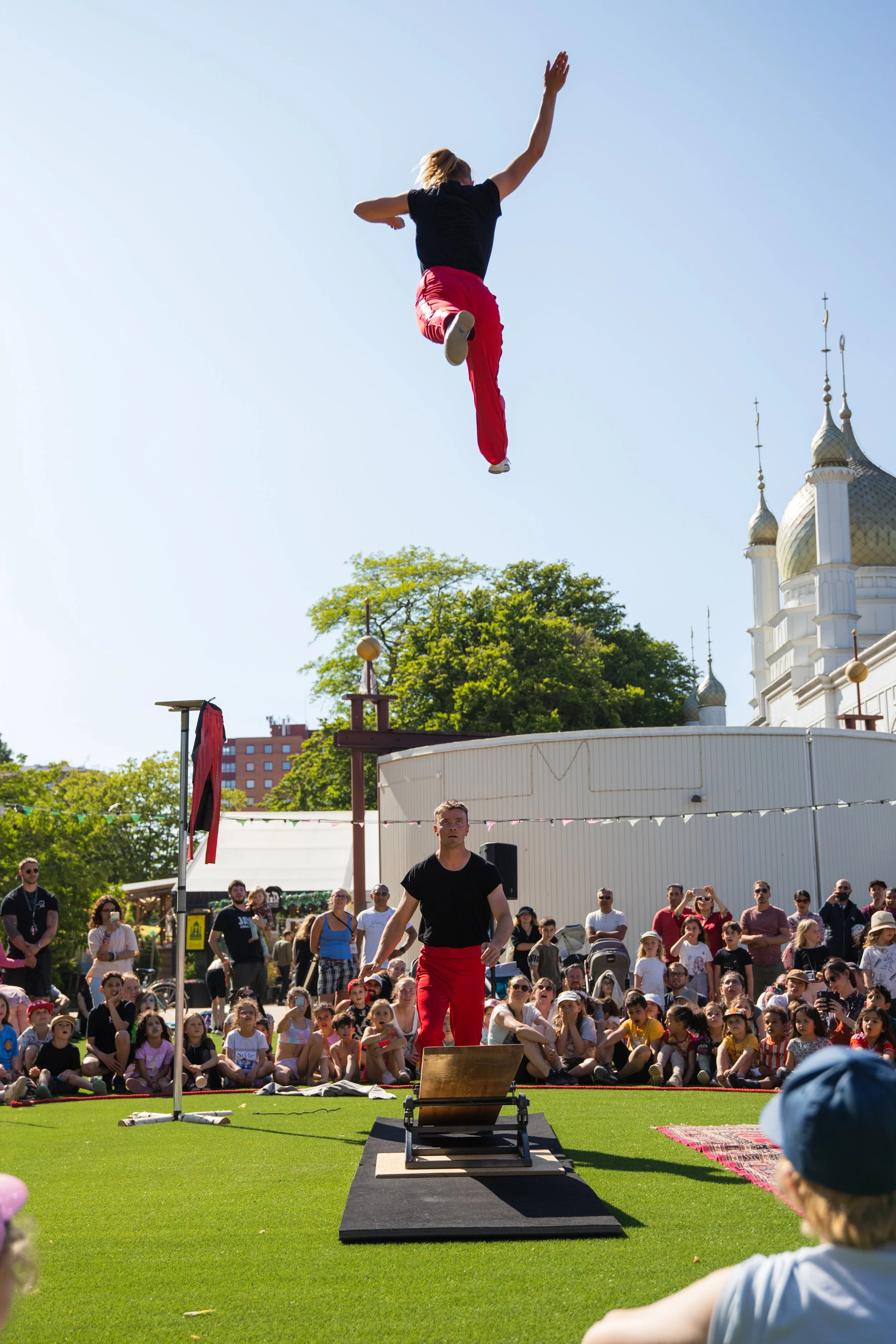 A performer in a black shirt and red pants is mid-air during a display at an outdoor event, with a crowd of children and adults watching on. The scene takes place on a sunny day with clear blue skies, green trees, and a building with domed rooftops i