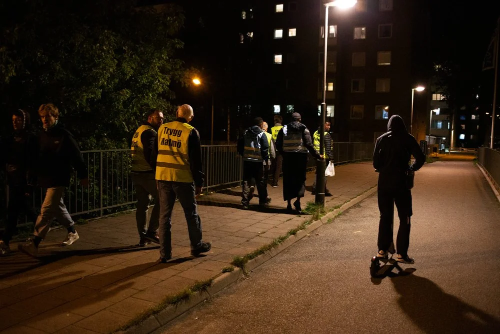 Group of people walking on a street at night with one person on a hoverboard, some wearing reflective vests.