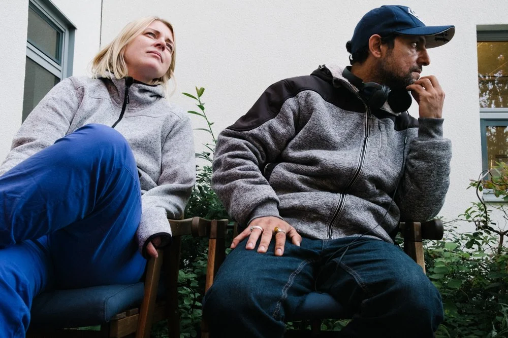 A woman and a man sitting outdoors on chairs, with the woman looking away pensively and the man wearing headphones, appearing thoughtful.