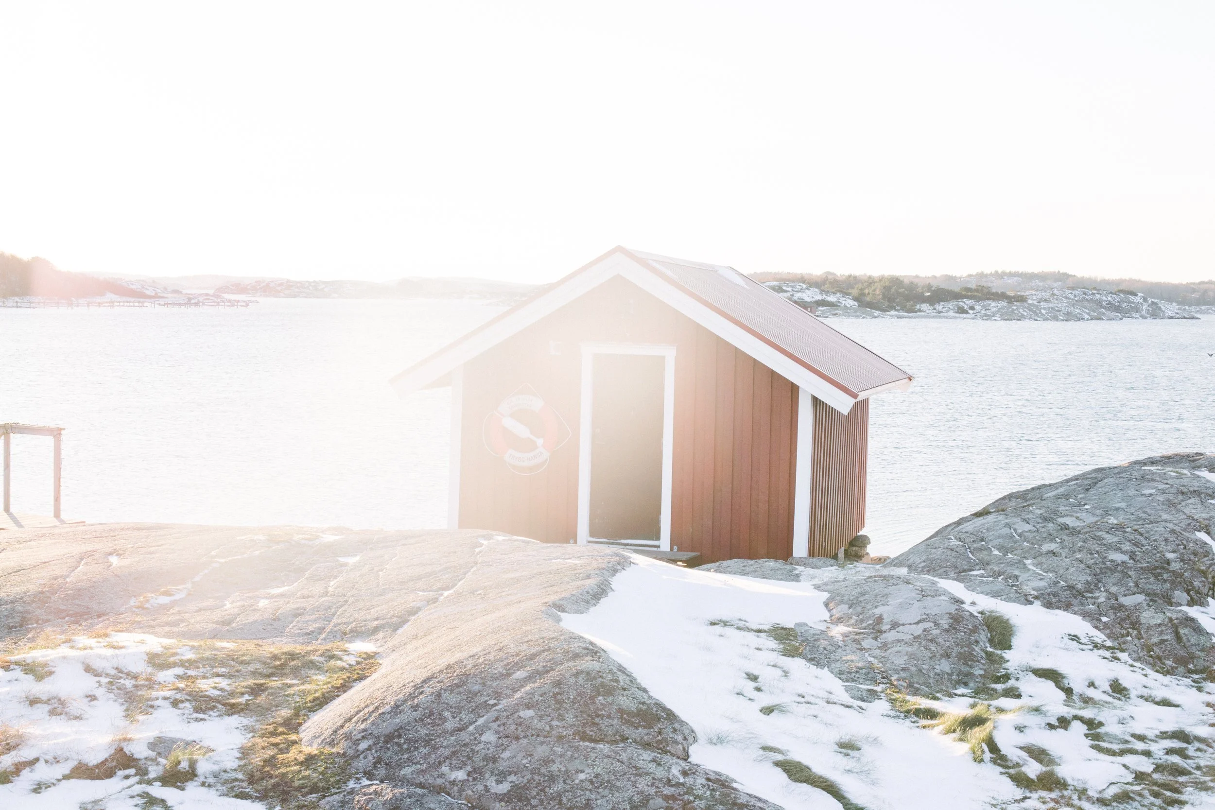 A small red wooden cabin with white trim on rocky, snow-covered terrain near a body of water, with snow-capped hills in the background and bright sunlight creating a hazy effect.