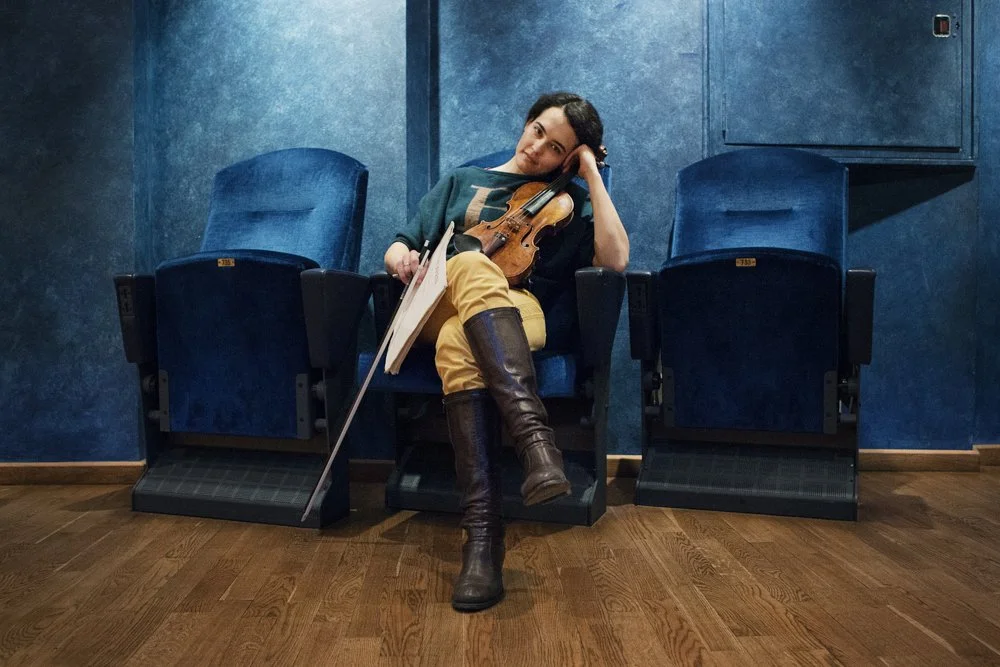 A woman sitting in a theater seat, resting her head on her hand, holding a violin and a bow. She is wearing a green shirt, yellow pants, and tall brown boots. There are two empty theater seats beside her, and the room has blue walls and wooden floori