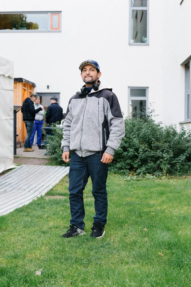 A man standing outdoors on a grassy area with a building in the background, wearing a gray and black jacket, jeans, a baseball cap, and headphones around his neck. Several people are in the background near a wooden structure.