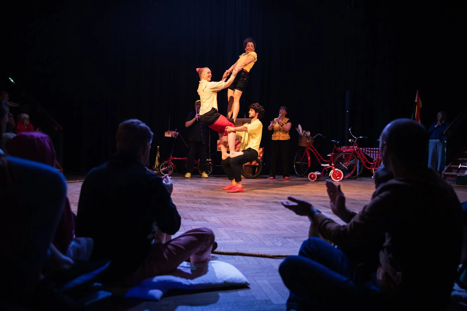 A circus performance featuring three performers in yellow shirts balancing in a human pyramid on stage, with an audience watching from the foreground and a band with musicians playing instruments in the background.