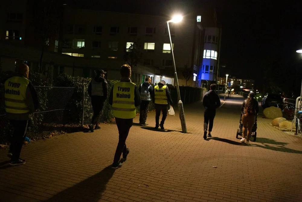 Nighttime scene of people walking on a lit city sidewalk, some wearing yellow vests with "Trygg Malmö" written on them, near residential buildings and large rocks.