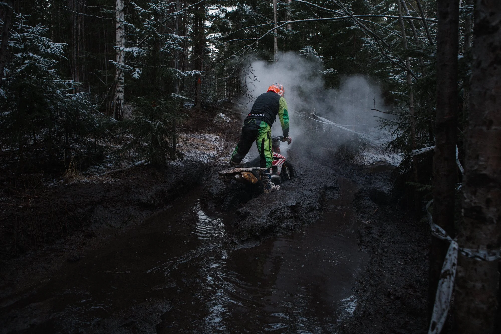 A person dressed in motocross gear, including a helmet and racing suit, standing on a muddy trail in a forest, with steam or smoke rising in the background.