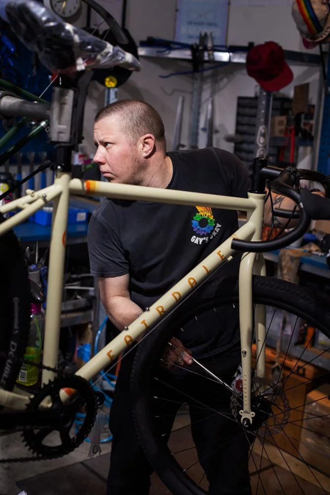 A man inspects a bicycle in a workshop, surrounded by tools and bike parts.