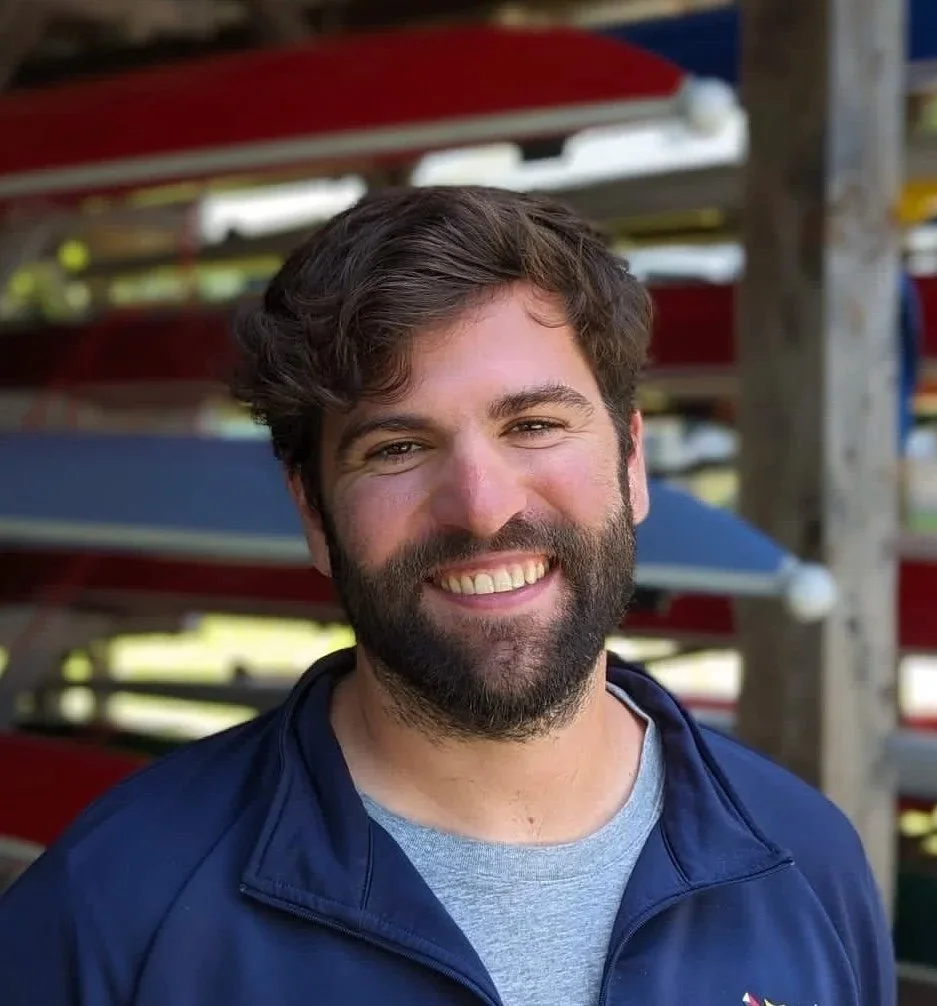 Smiling man with a beard and wavy hair, wearing a navy jacket over a gray T-shirt, standing outdoors near a rack of colorful kayaks.