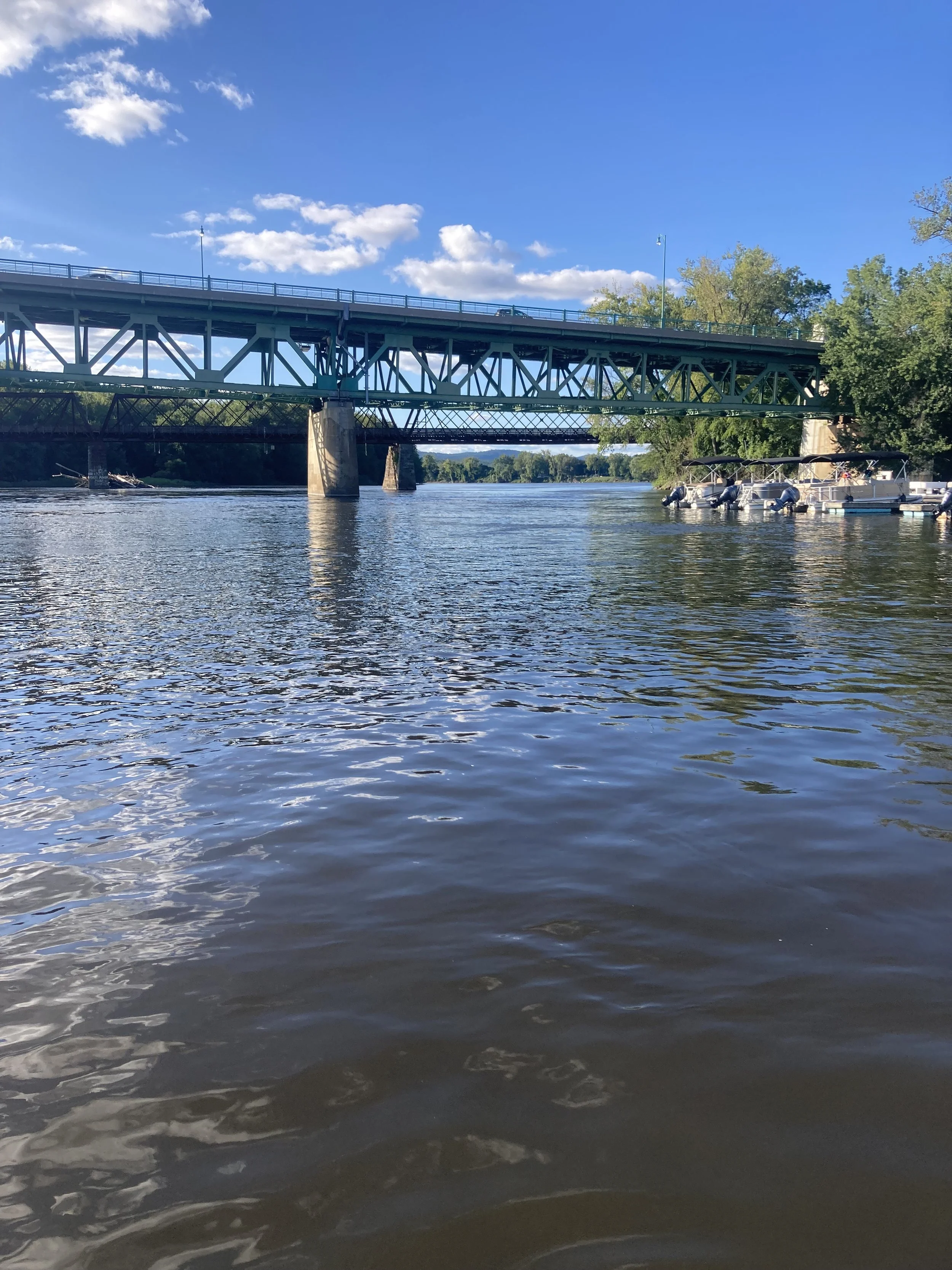 A river scene with a green metal bridge overhead, boats docked on the right, trees along the banks, and a partly cloudy blue sky.