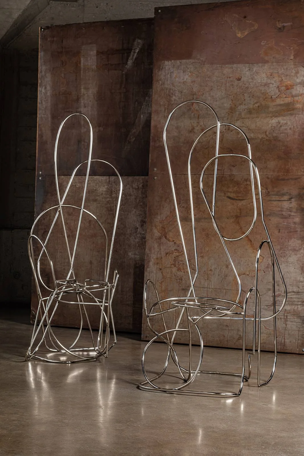Two modern abstract sculptures made of shiny metal wire, resembling chairs or figures, set against a rusty, aged metal background.