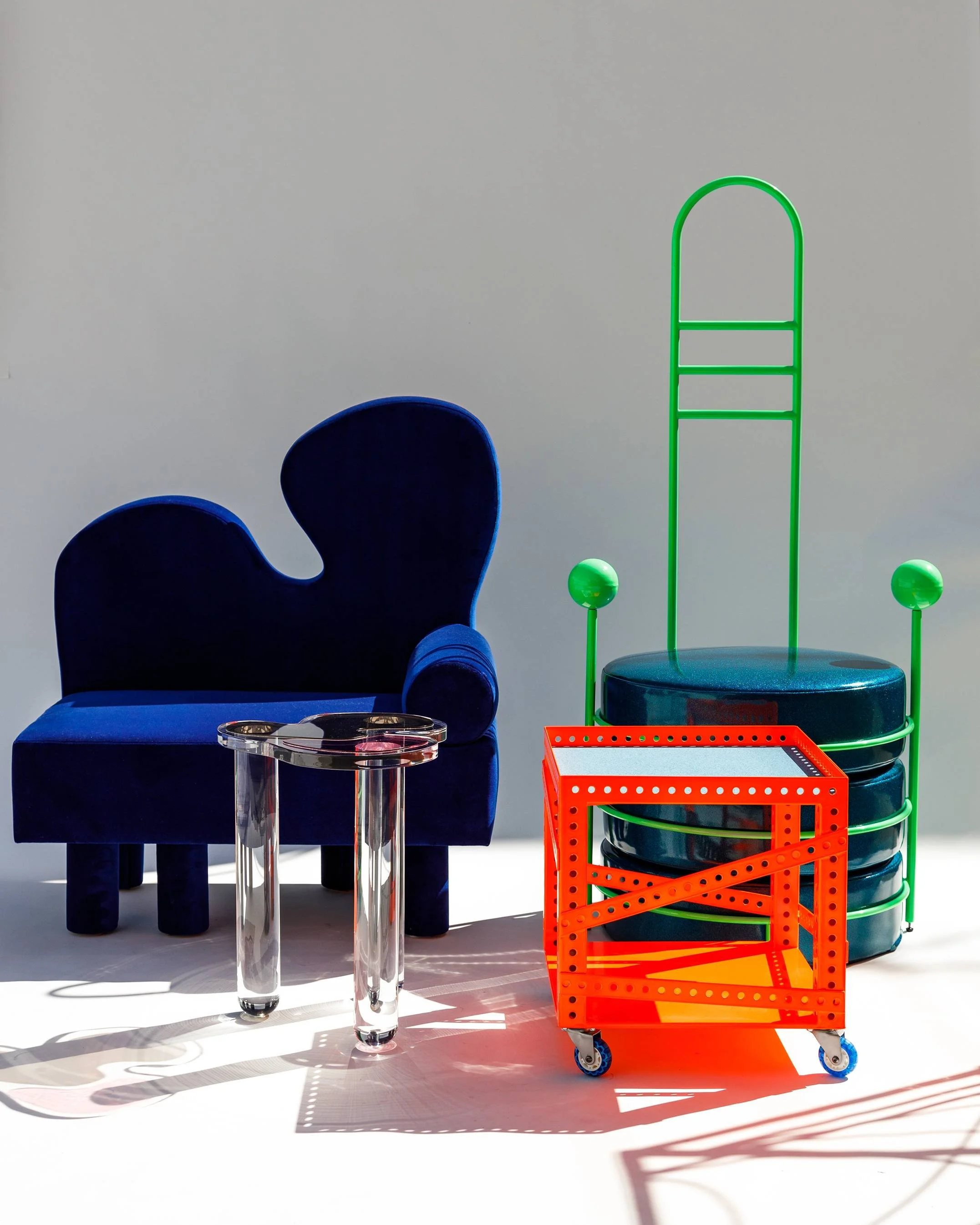 Assorted modern furniture and decor items including a blue velvet armchair, a small transparent side table, a green metal chair with a round blue seat, and a red rolling cart, set against a plain light background.