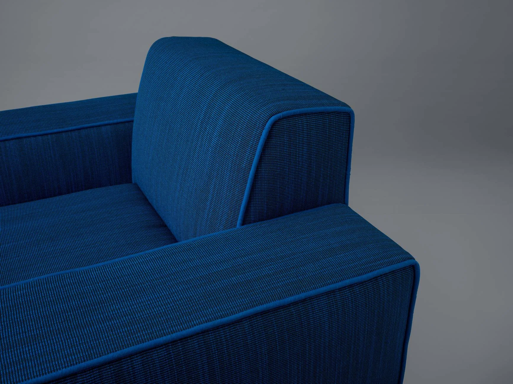 Close-up of a modern blue upholstered sofa with textured fabric and piping details against a plain gray background.