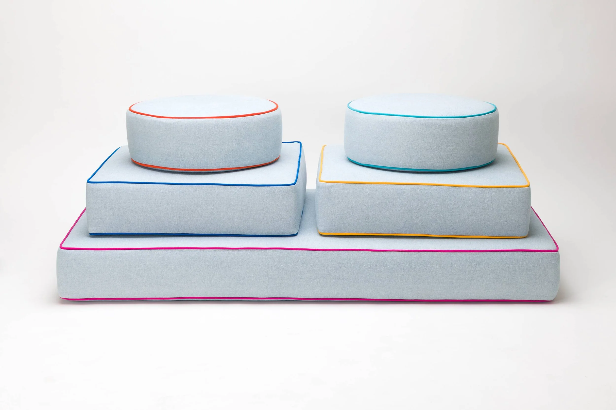 Stacked modern, colorful upholstered cushions with spiral edges in pink, blue, yellow, and orange on a plain white background.