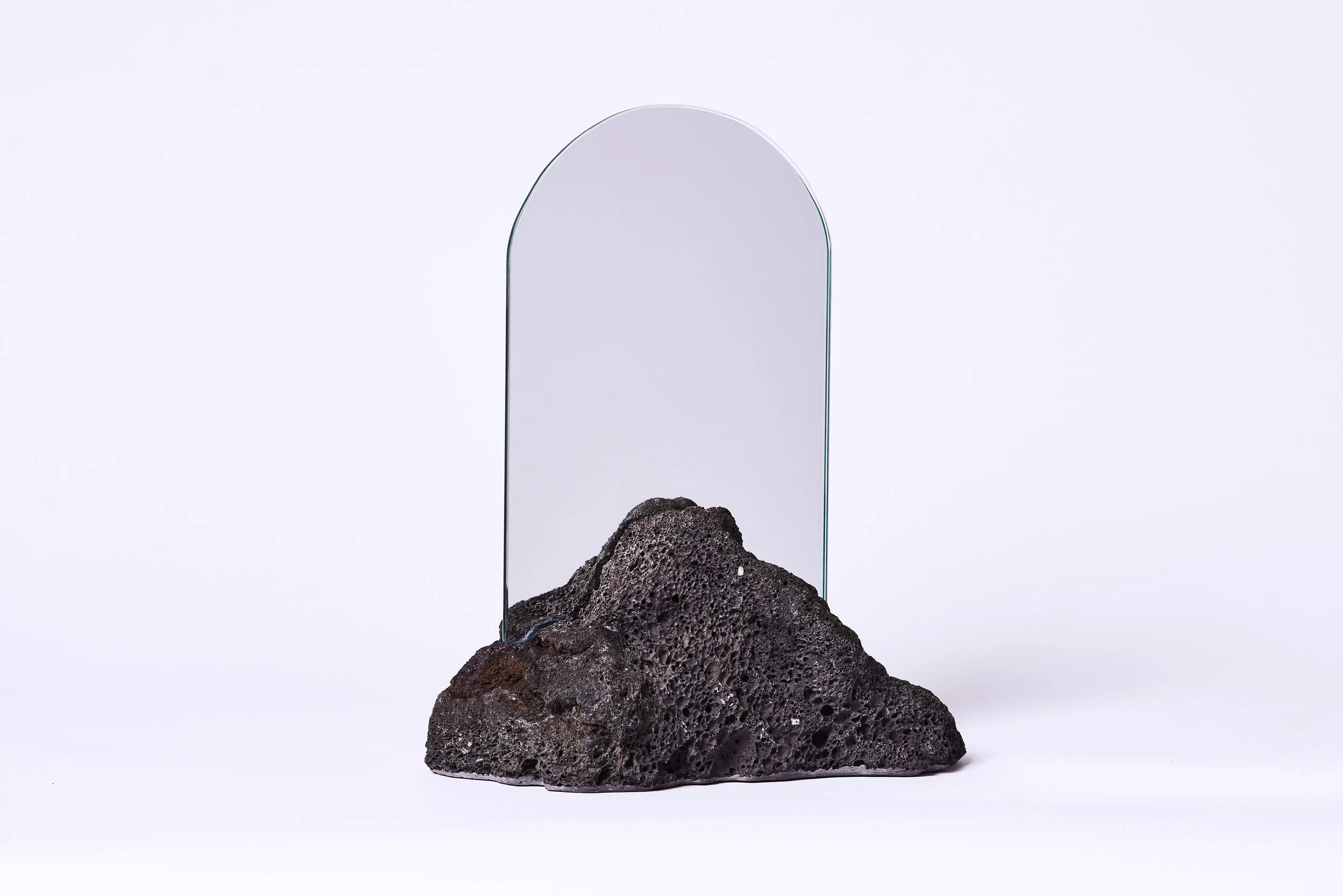A black volcanic rock with a rough, porous surface, placed in front of a tall, arched mirror on a white background.