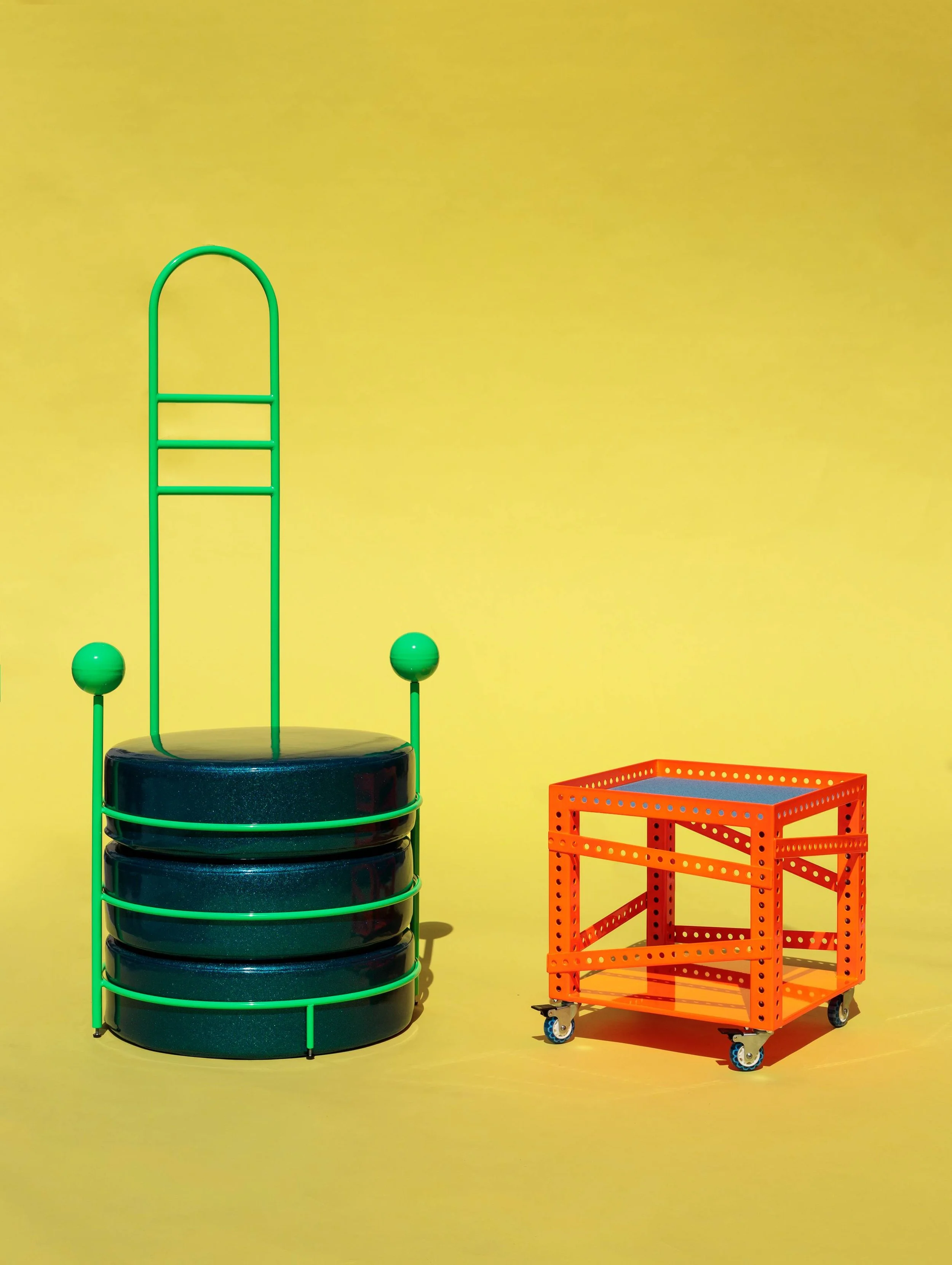 Colorful modern furniture pieces including a green metal chair with a round black seat and a matching green backrest, and an orange rolling cart with a square frame and perforated sides, set against a yellow background.