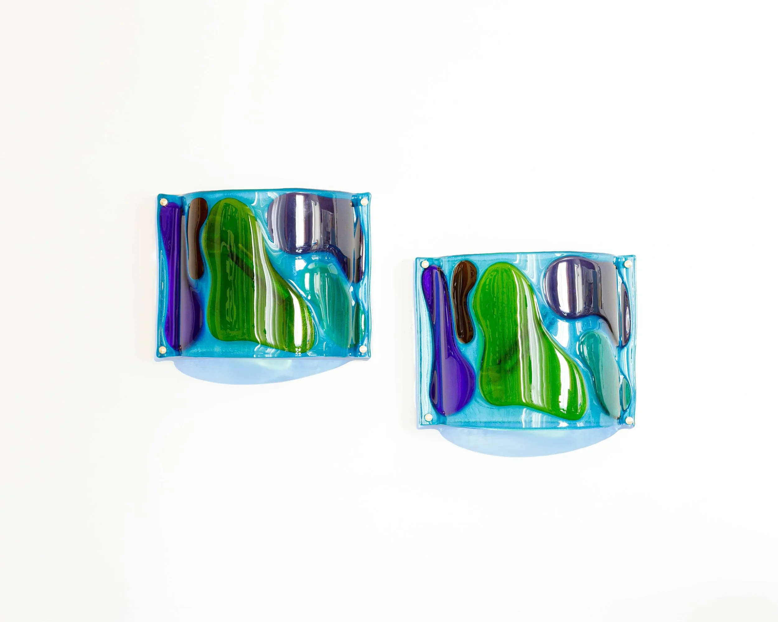 Two square glass art pieces with abstract blue, green, purple, and brown designs hanging on a white wall.