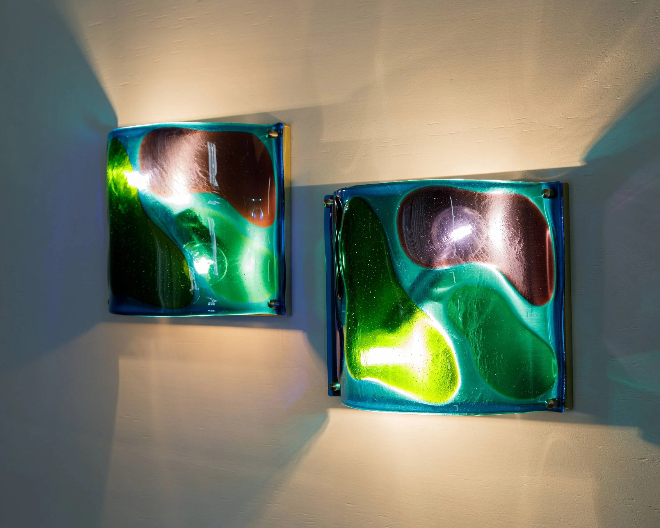 Two abstract glass wall art pieces with swirling green, blue, and brown colors, illuminated from behind, mounted on a beige wall.