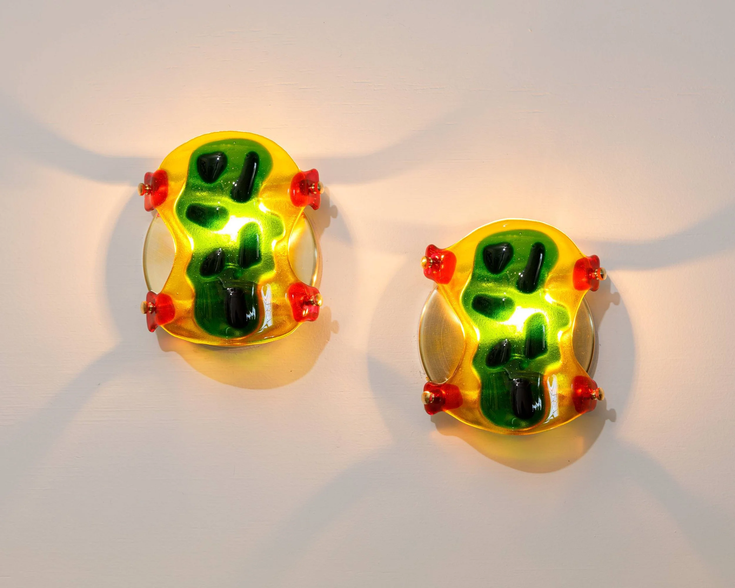Two colorful, abstract glass sculptures with yellow, green, and black colors, each mounted on a wall with shadows cast below.