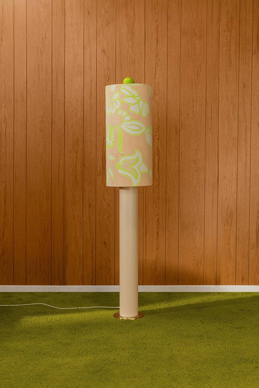 A floor lamp with a floral-patterned shade on a wood-paneled wall and green carpet floor.