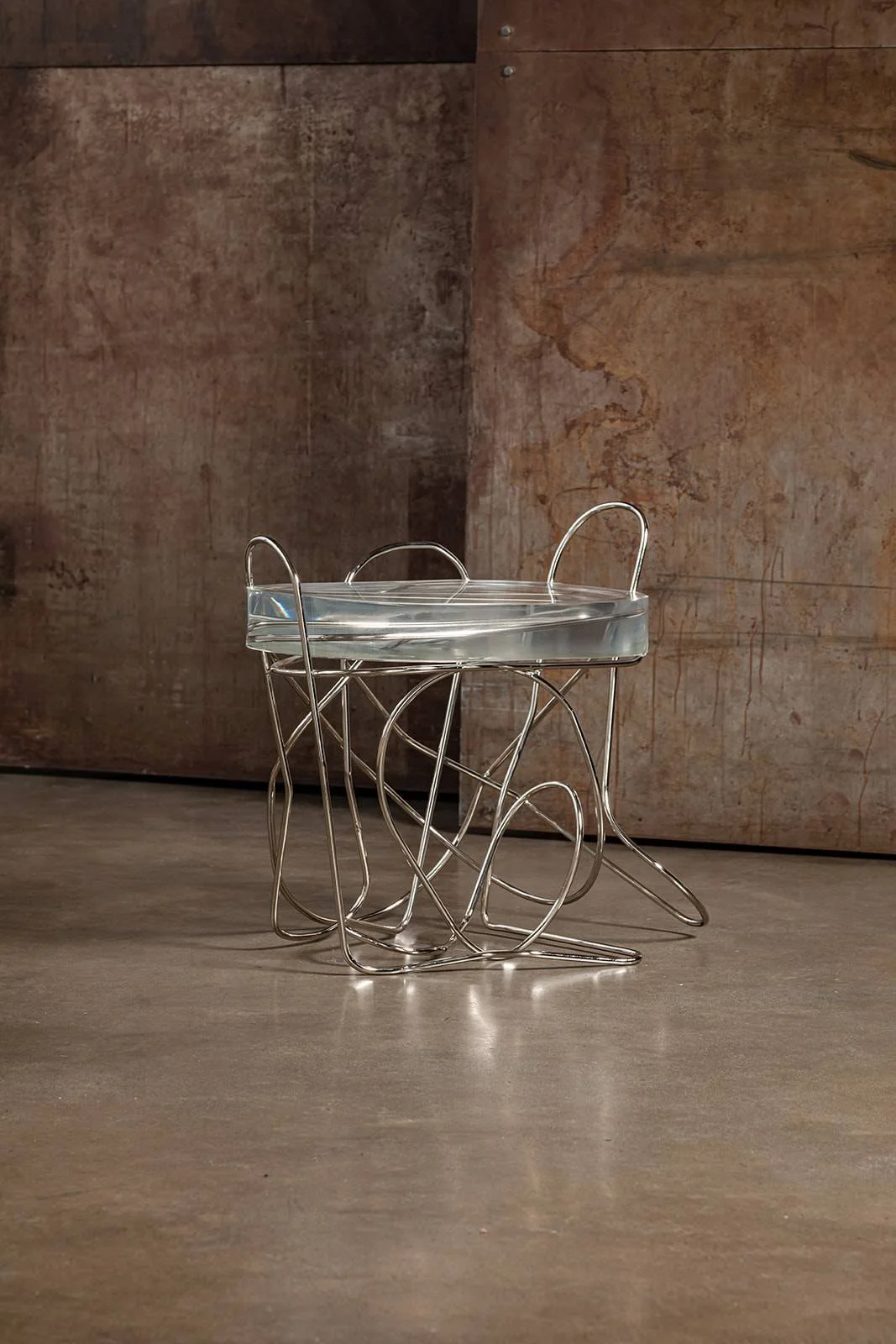 A modern glass-top table with an intricate silver metal wire base, placed on a polished concrete floor against a textured brown wall with subtle markings.