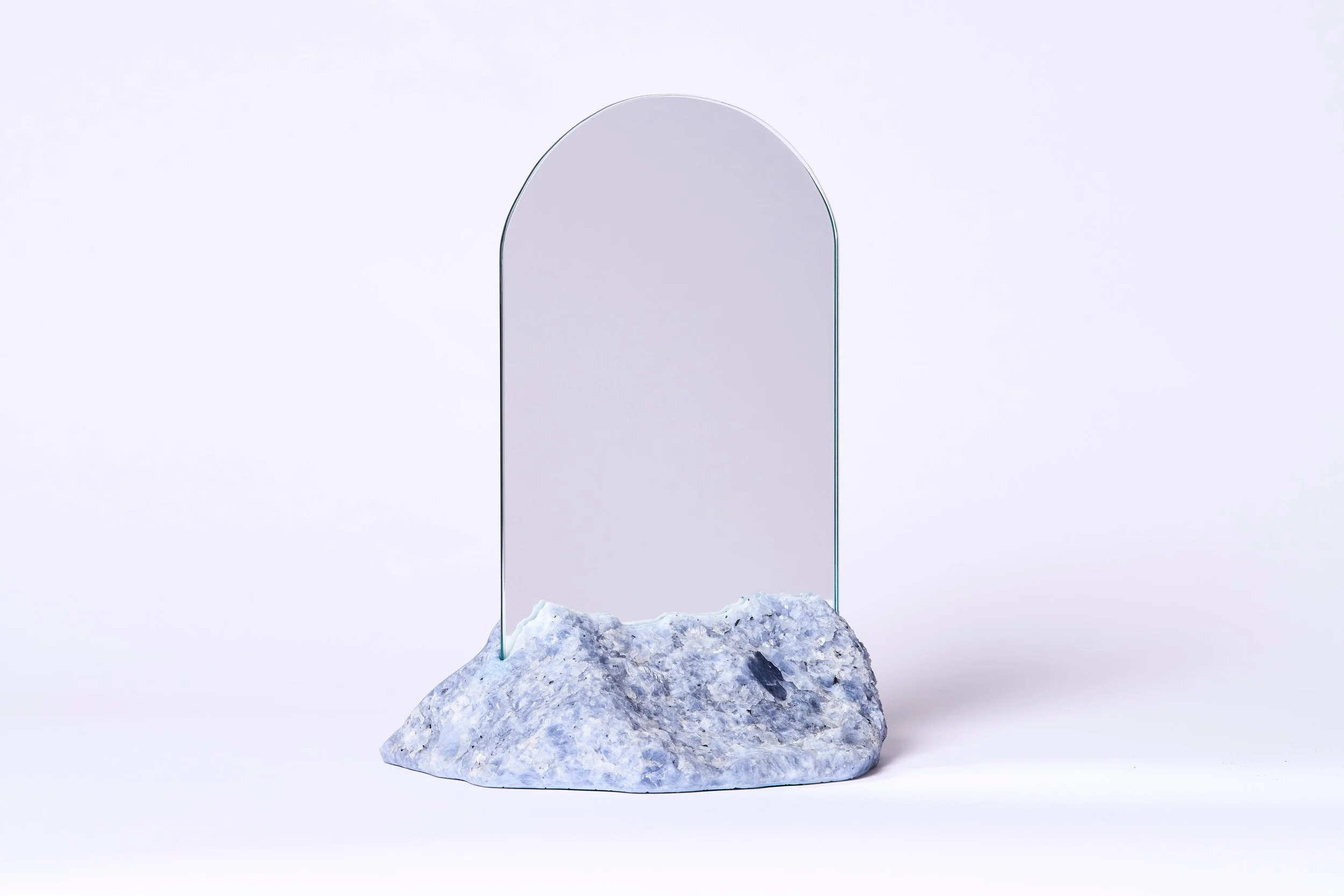 A mirror with a rounded top sits on a rocky base against a plain white background.