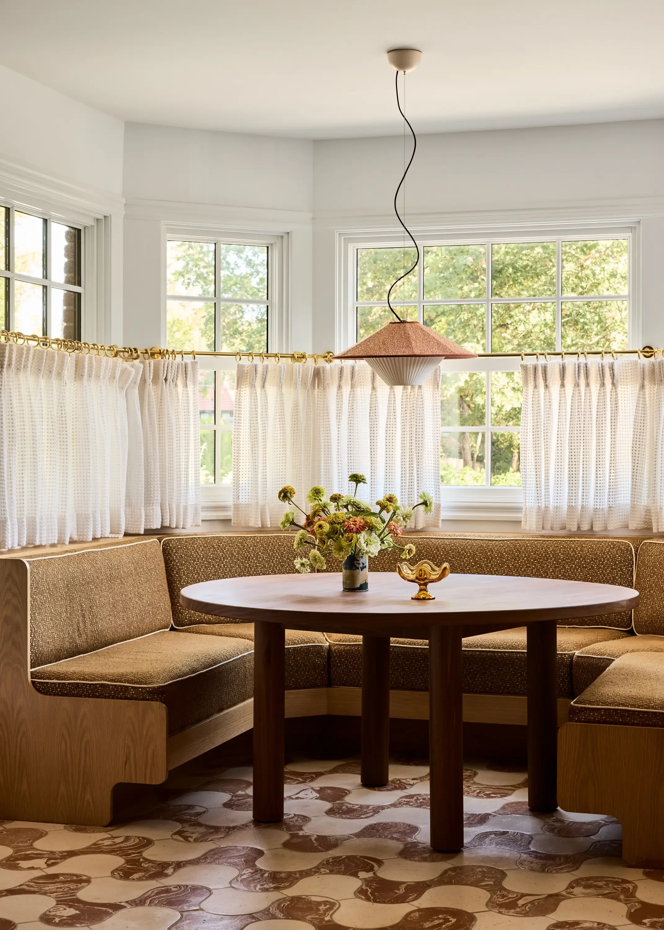 Vintage kitchen nook with a rounded wooden table, floral centerpiece, and patterned floor tiles, surrounded by a built-in upholstered bench, with lots of natural light and white curtains.