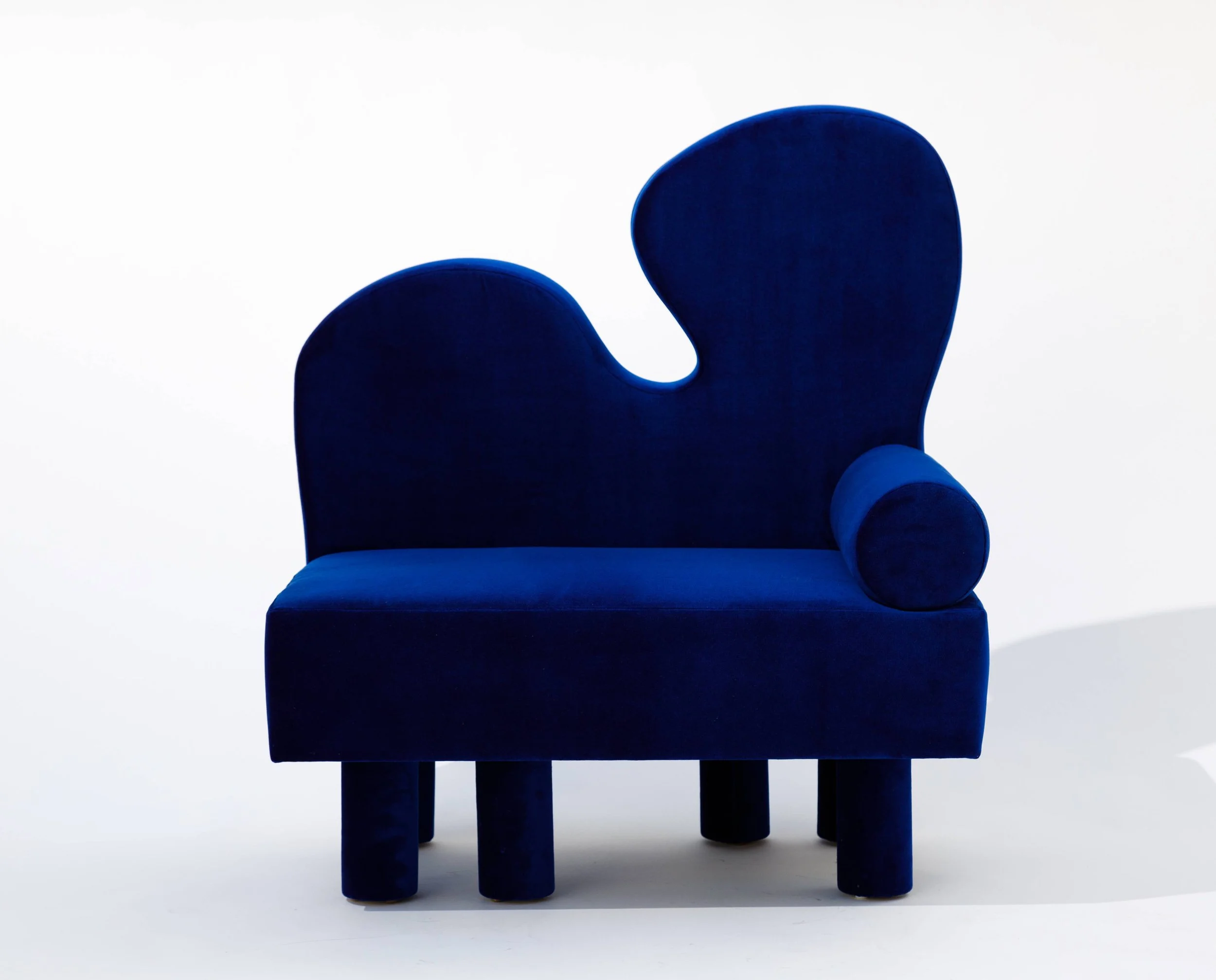 A modern, artistic blue chair with a rounded backrest, curved armrest, and four cylindrical legs, set against a plain white background.
