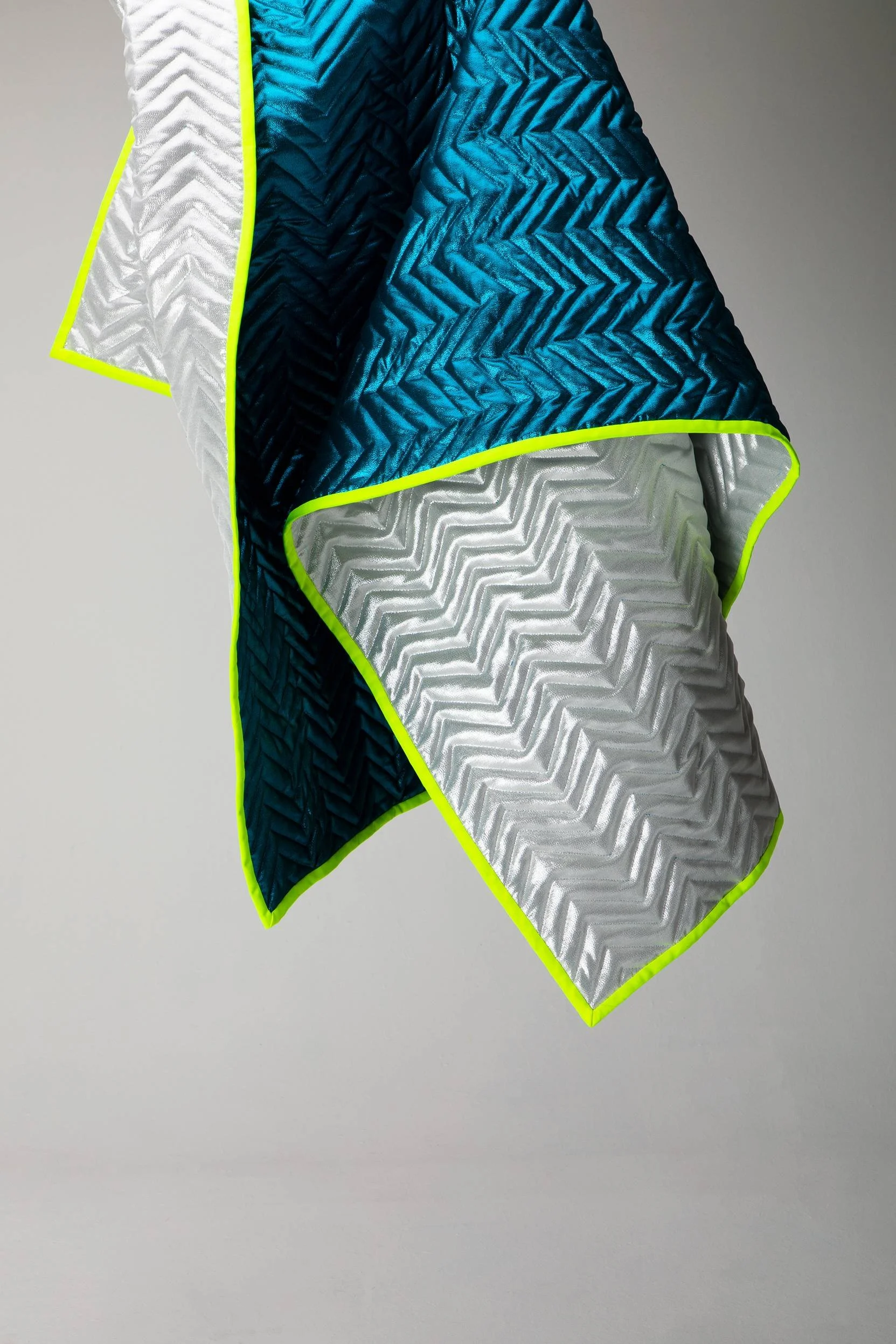 Close-up of two quilted fabric pieces, one in bright blue and the other in silver, with neon yellow trim, draped against a plain gray background.