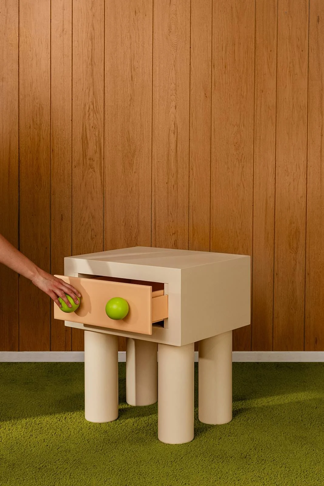 A small, modern white bedside table with a drawer that has peach-colored knobs, placed on a green carpet against a wood-paneled wall. A person's hand is opening the drawer and holding a small green ball.