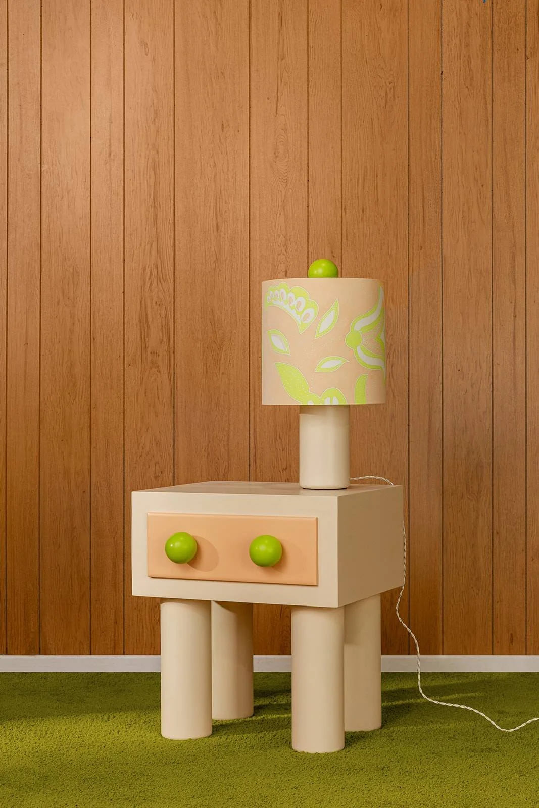 A small Tan and Peach-colored side table with two round green knobs on the drawer, standing on four cylindrical legs. On top of the table is a small lamp with a white base and a shade with green leaf and flower patterns, topped with a green ball. The