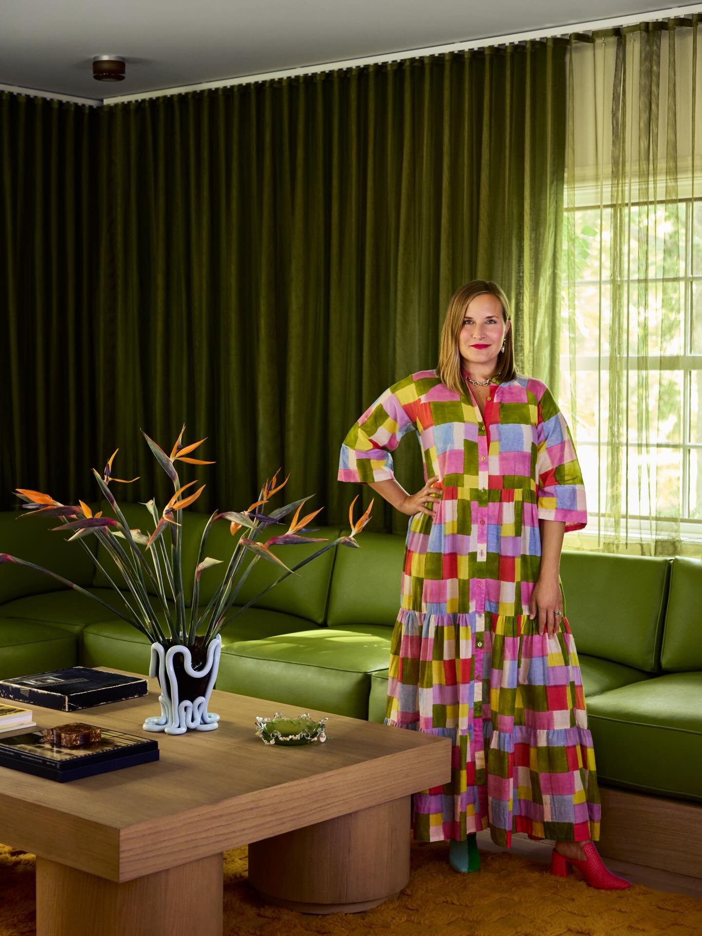 Woman standing in a living room with green curtains, a green sectional sofa, a wooden coffee table with books, a decorative vase with flowers, and a window letting in sunlight. She is wearing a colorful, checkered dress and pink shoes.