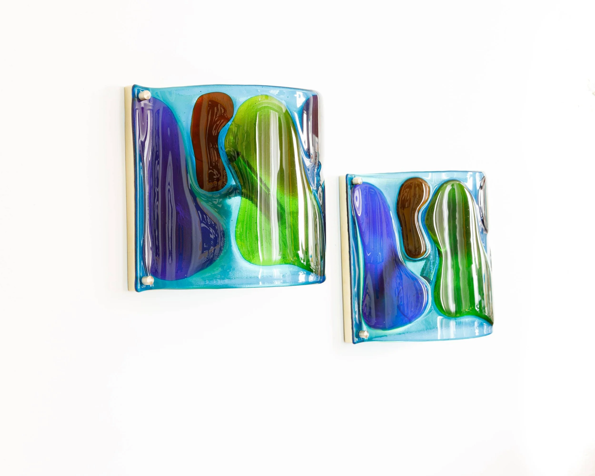 Two square glass art pieces with colorful, abstract swirling patterns in blue, green, brown, and purple, mounted on a white wall.