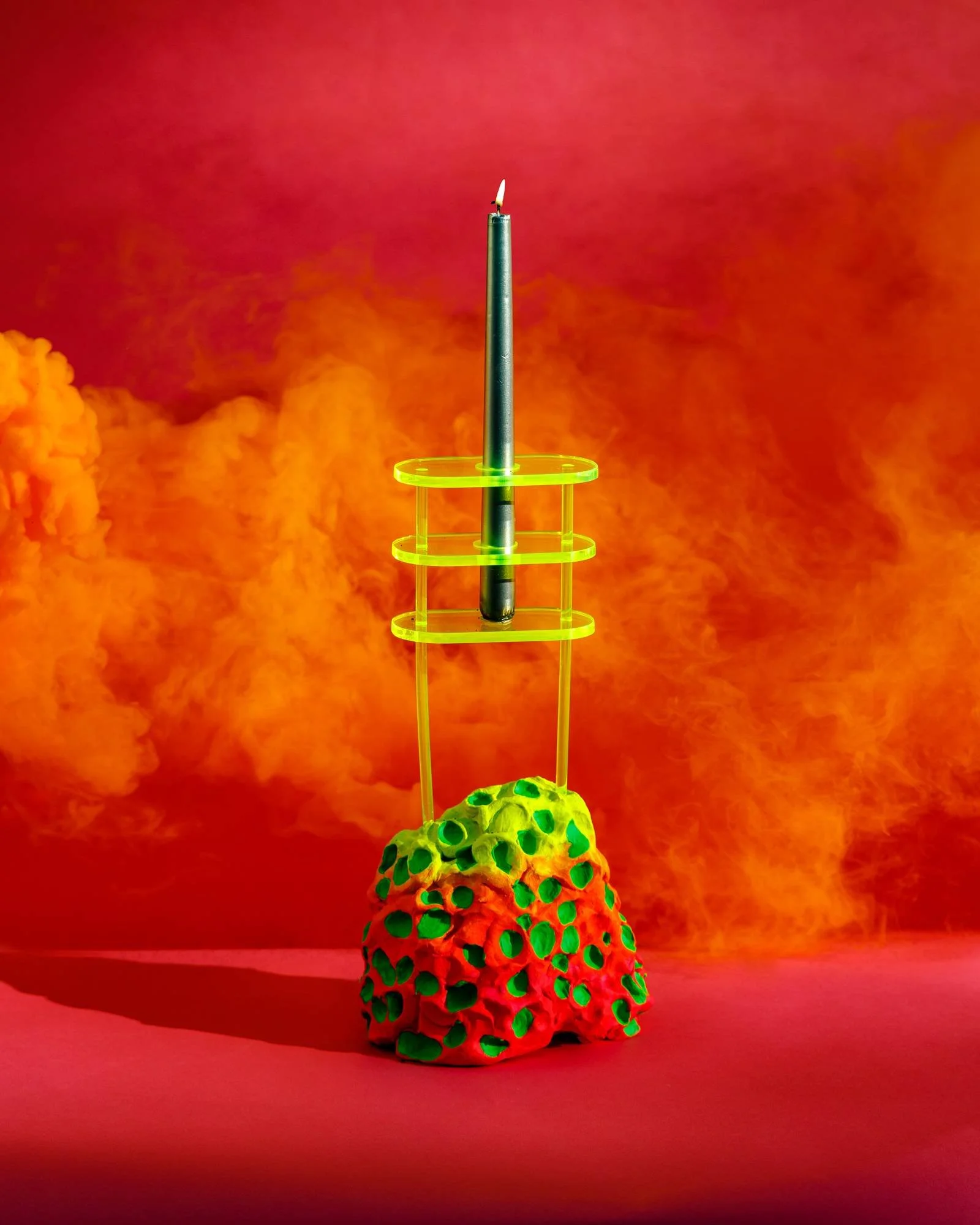 A lit green candle on top of a neon green wire structure, perched on a multicolored porous base with holes, against an orange and pink smoky background.