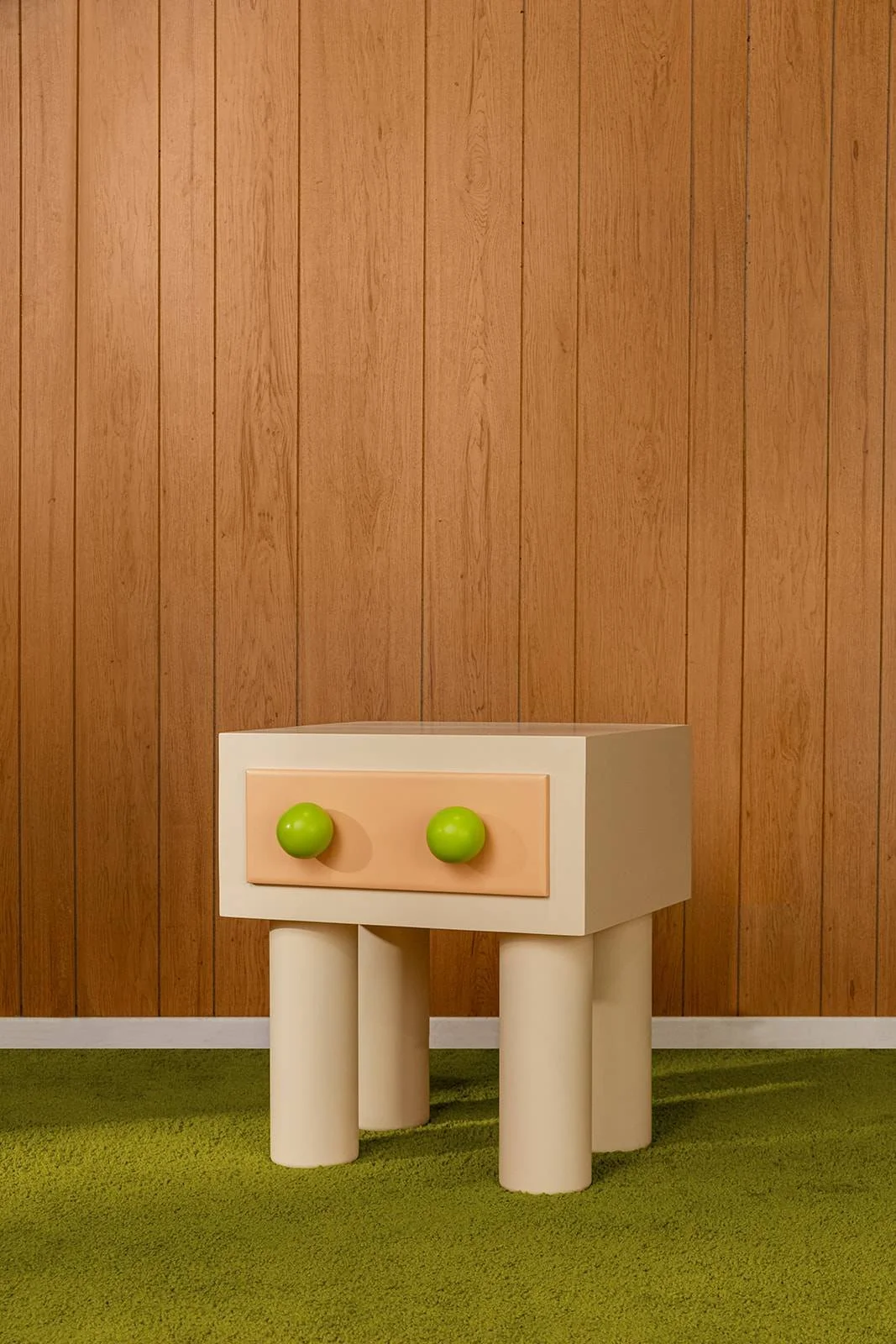A small white nightstand with two green knobs, standing on four short white legs, against a wood-paneled wall on a green carpeted floor.