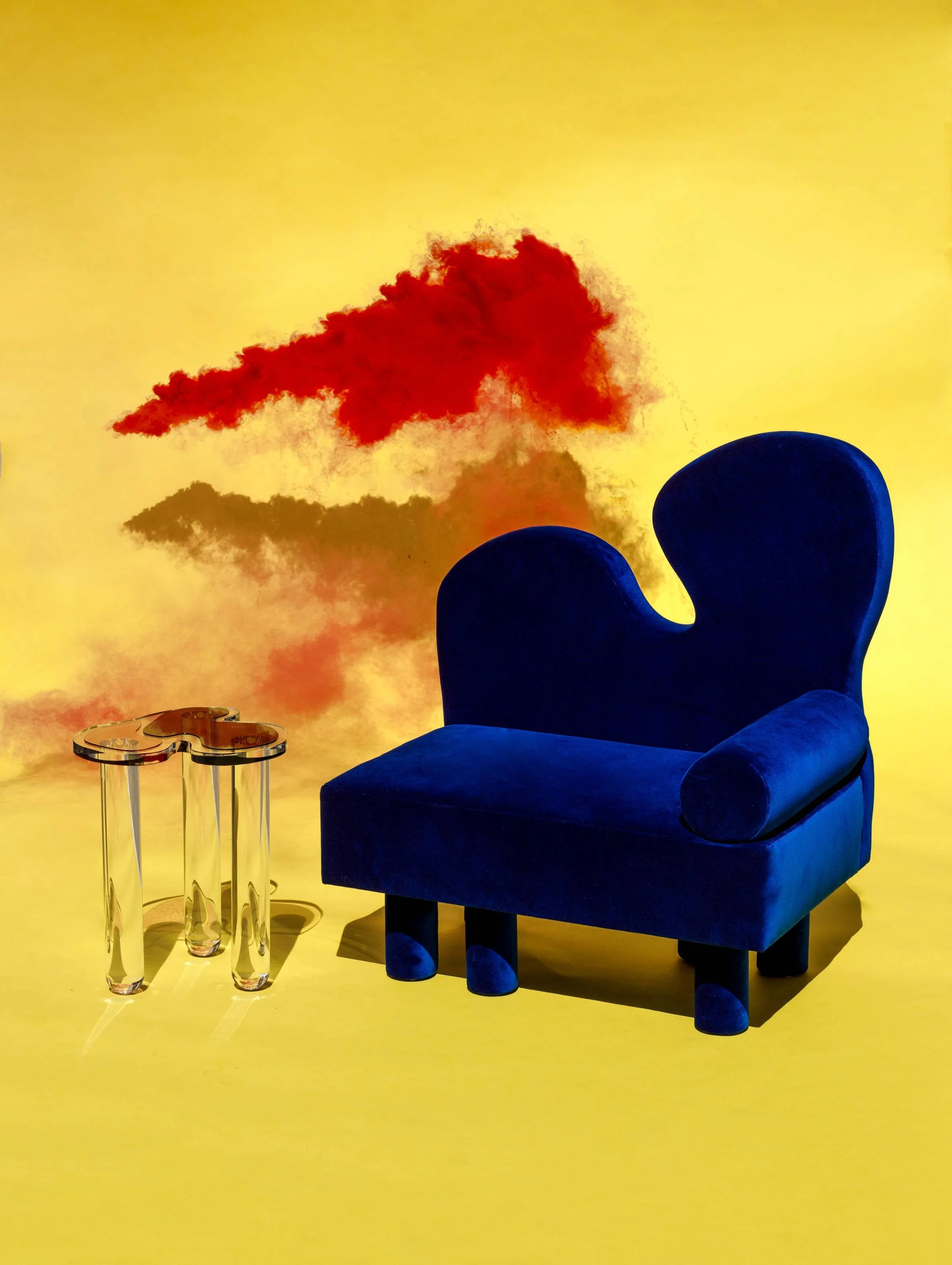 A blue velvet chair with a rounded backrest, a matching cylindrical cushion, and short legs, placed on a yellow floor. Next to the chair is a small transparent side table with a gold-colored top, featuring a decorative design. The background is a yel