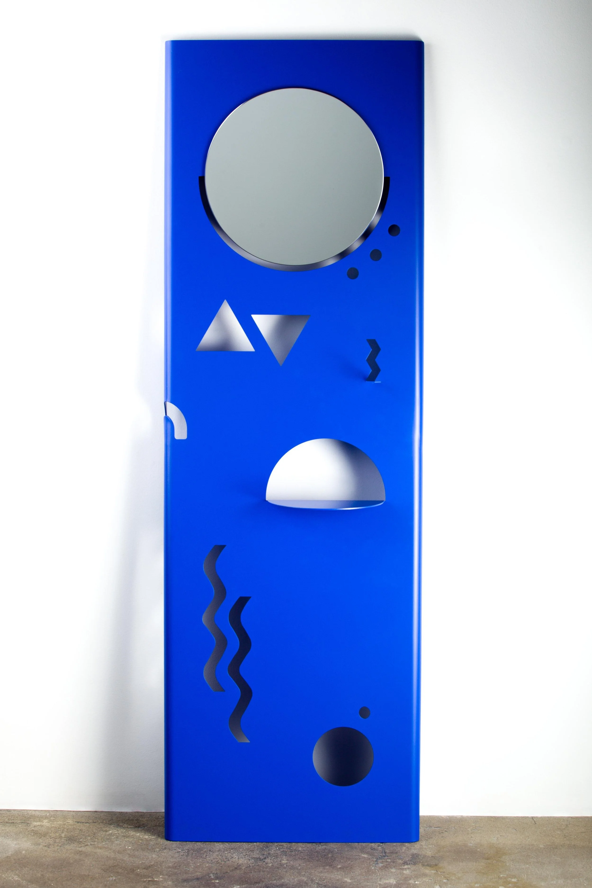 Blue modern sculpture with geometric cutouts and circular openings standing on concrete floor against white wall.