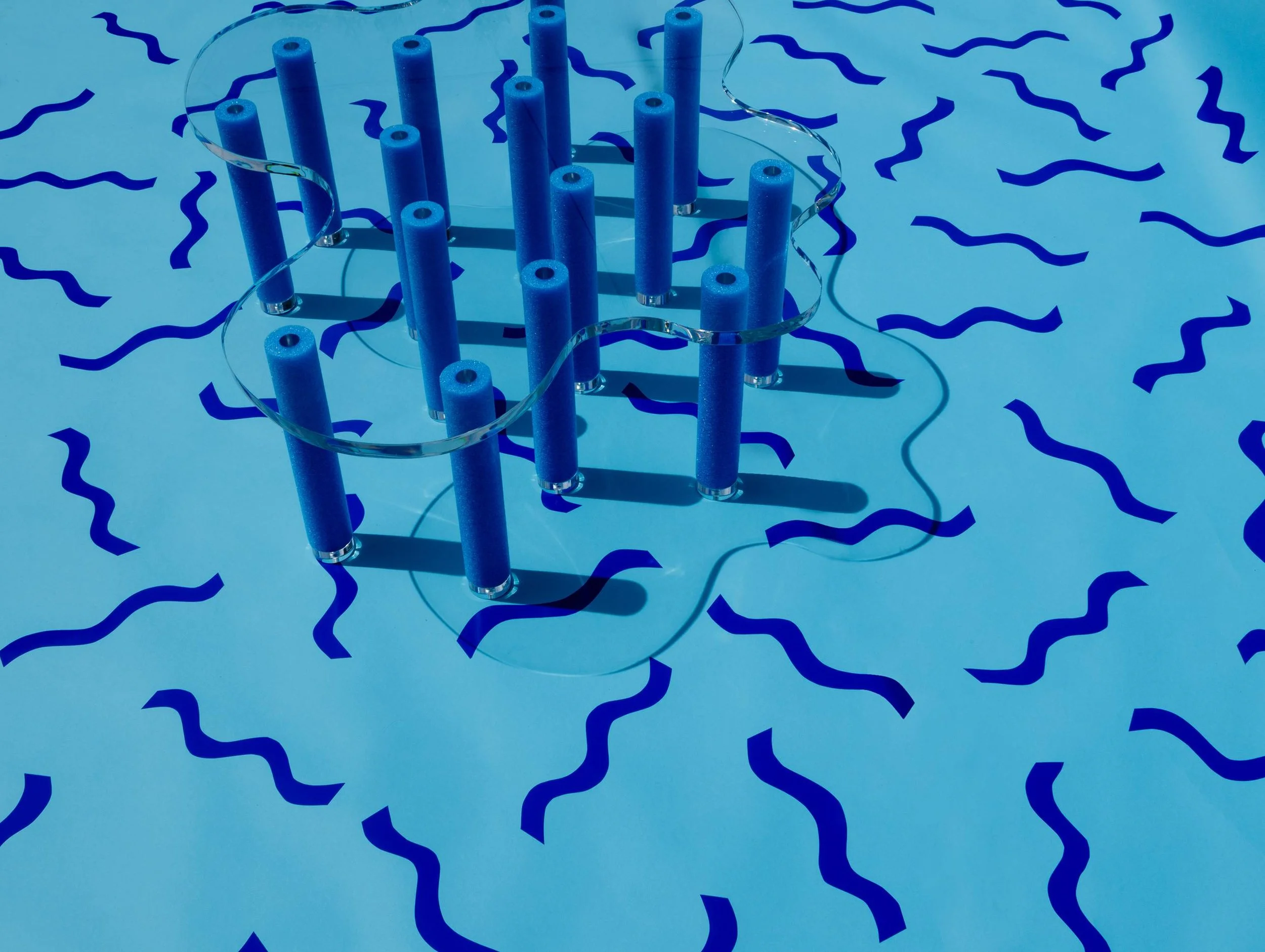 A blue abstract sculpture made of vertical cylindrical rods connected by a clear curved strip, placed on a light blue surface with darker blue wavy lines.