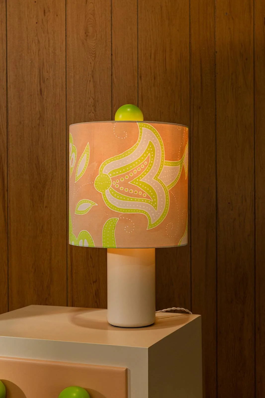 Table lamp with a floral-patterned lampshade featuring pink and green flower design, green decorative ball on top, and a white base, placed on a beige surface against a wood-paneled wall background.