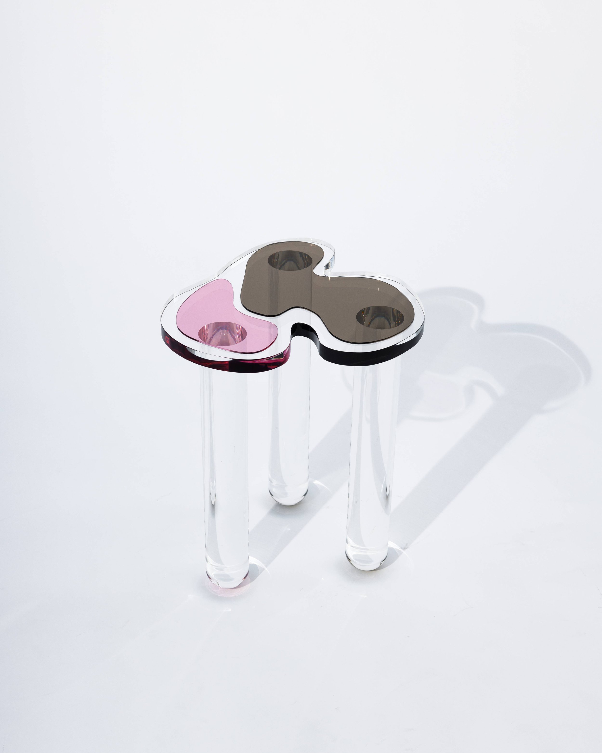 A modern artistic glass sculpture with an abstract butterfly shape, featuring pink and brown sections, supported by three clear acrylic legs.