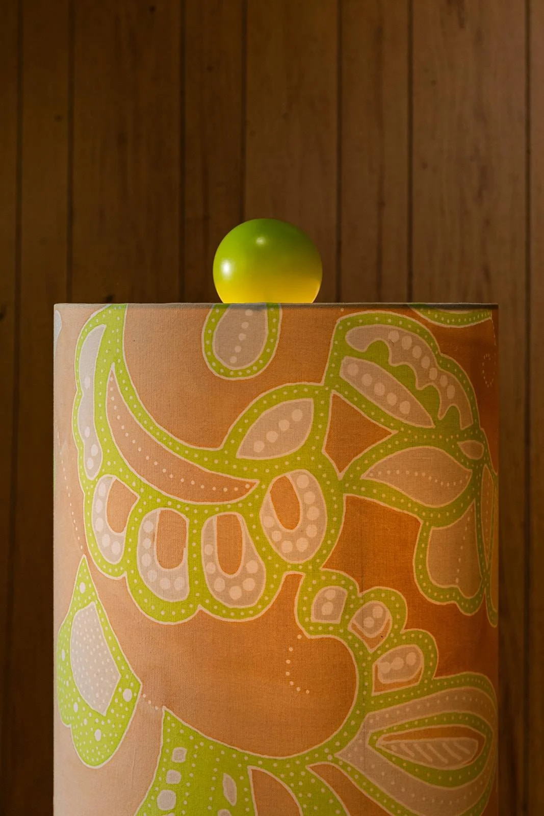 A green apple on top of a cylindrical object with a colorful floral and leaf pattern, against a wooden-paneled wall background.