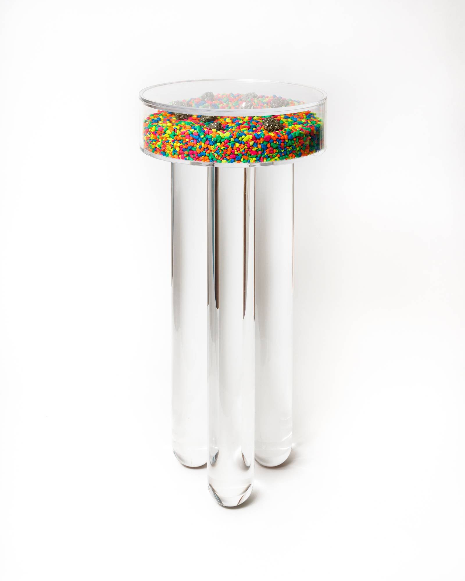 A clear, round glass container filled with colorful tiny sprinkles, sitting on top of three elongated clear glass tubes.