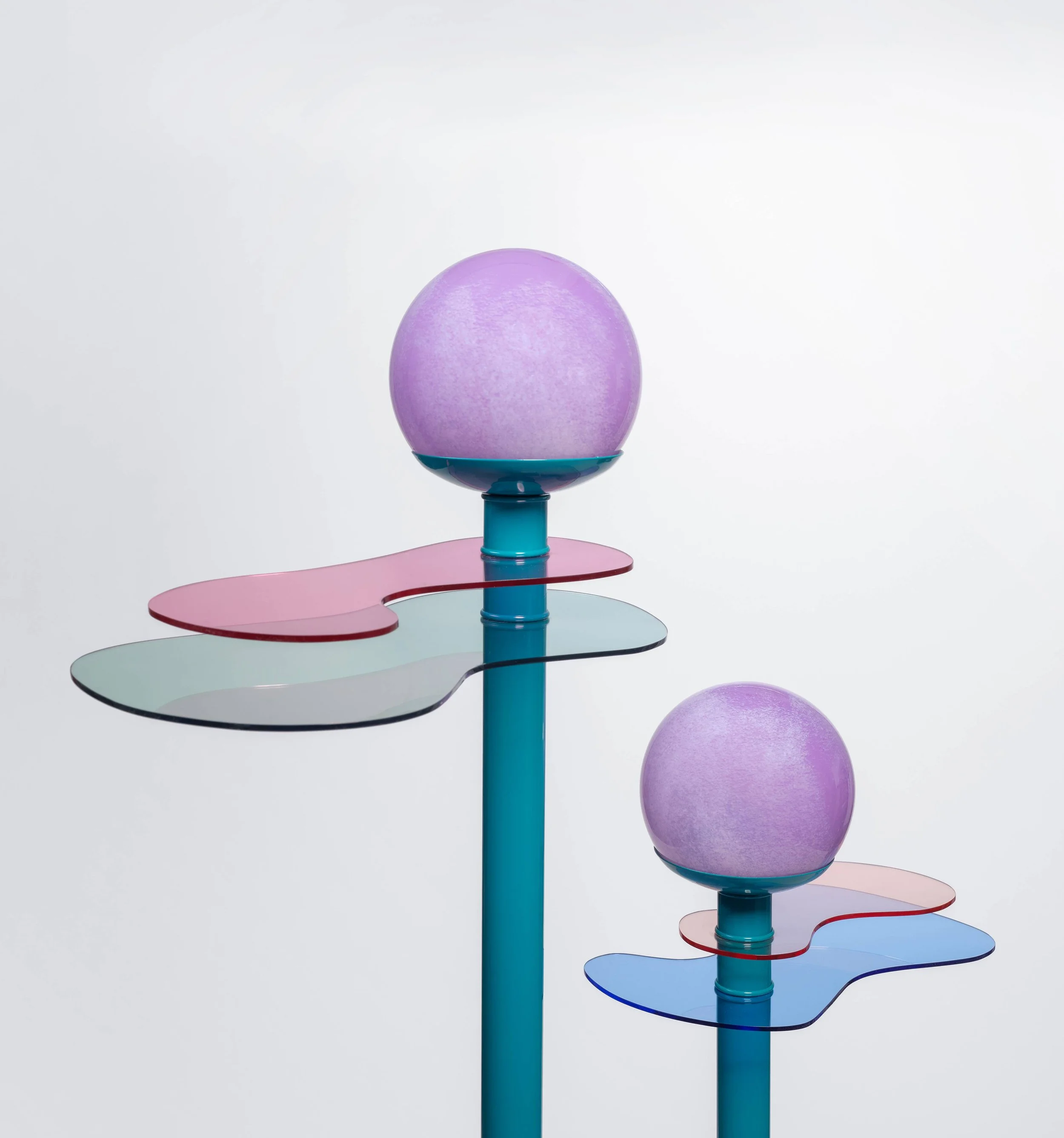 Two modern, colorful sculptures featuring tall blue poles with spherical purple orbs on top and curved, translucent platforms in pink, green, and blue.