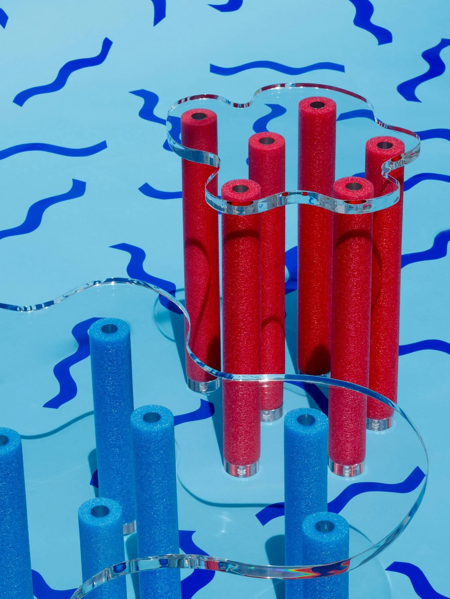 A decorative abstract art piece with vertical red and blue textured tubes on a light blue background, connected by transparent wavy acrylic strips.