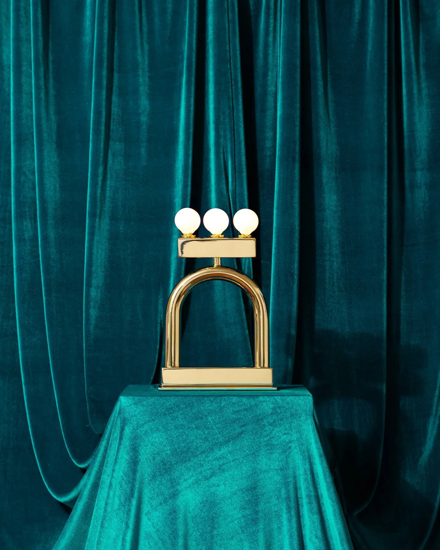 A modern decorative lamp with three white spherical bulbs on a gold and white base, placed on a teal velvet-covered table with matching teal velvet curtains in the background.