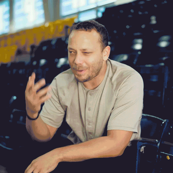 A man sitting in a stadium or auditorium, talking or explaining something with a smile, with empty black seats behind him.
