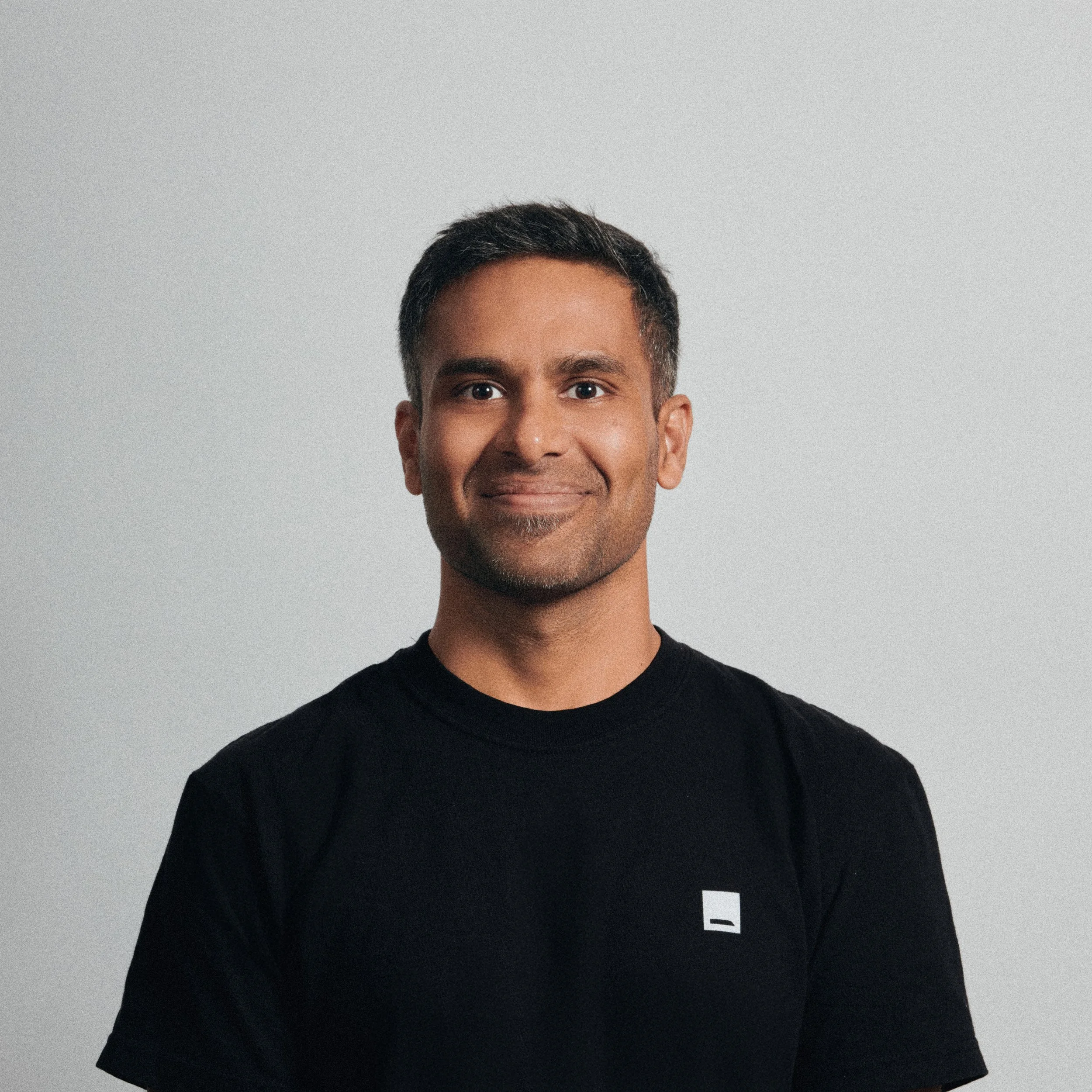 A man with short dark hair and a beard, wearing a black t-shirt with a small white logo on the chest, is standing against a plain light gray background.