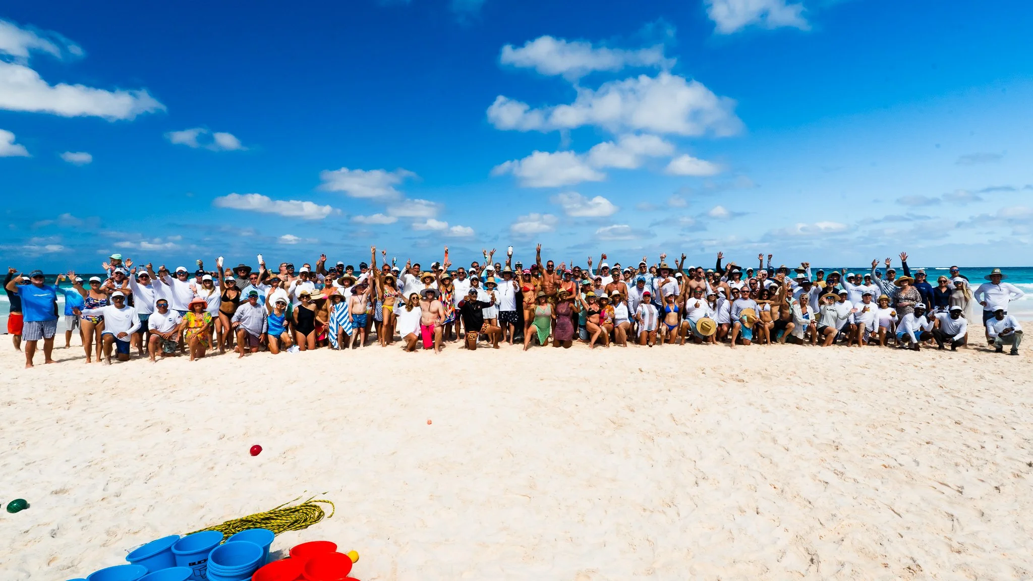 A large group of people on a sandy beach, dressed in swimsuits and summer clothing, facing the camera and enjoying a sunny day with blue skies and ocean in the background.